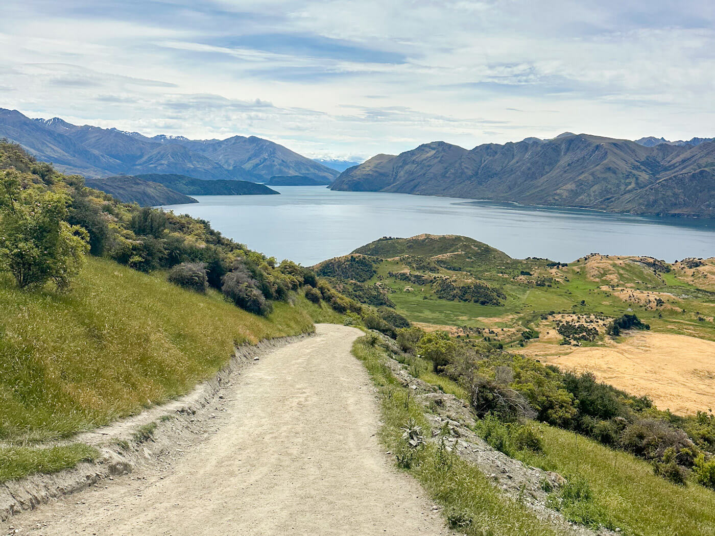 A wide, light-colored gravel path curves downward toward the center of the frame, flanked by green grass and low bushes. The view opens up to the blue lake and a series of smaller, green hills in the mid-ground, with high mountain peaks visible on the horizon.