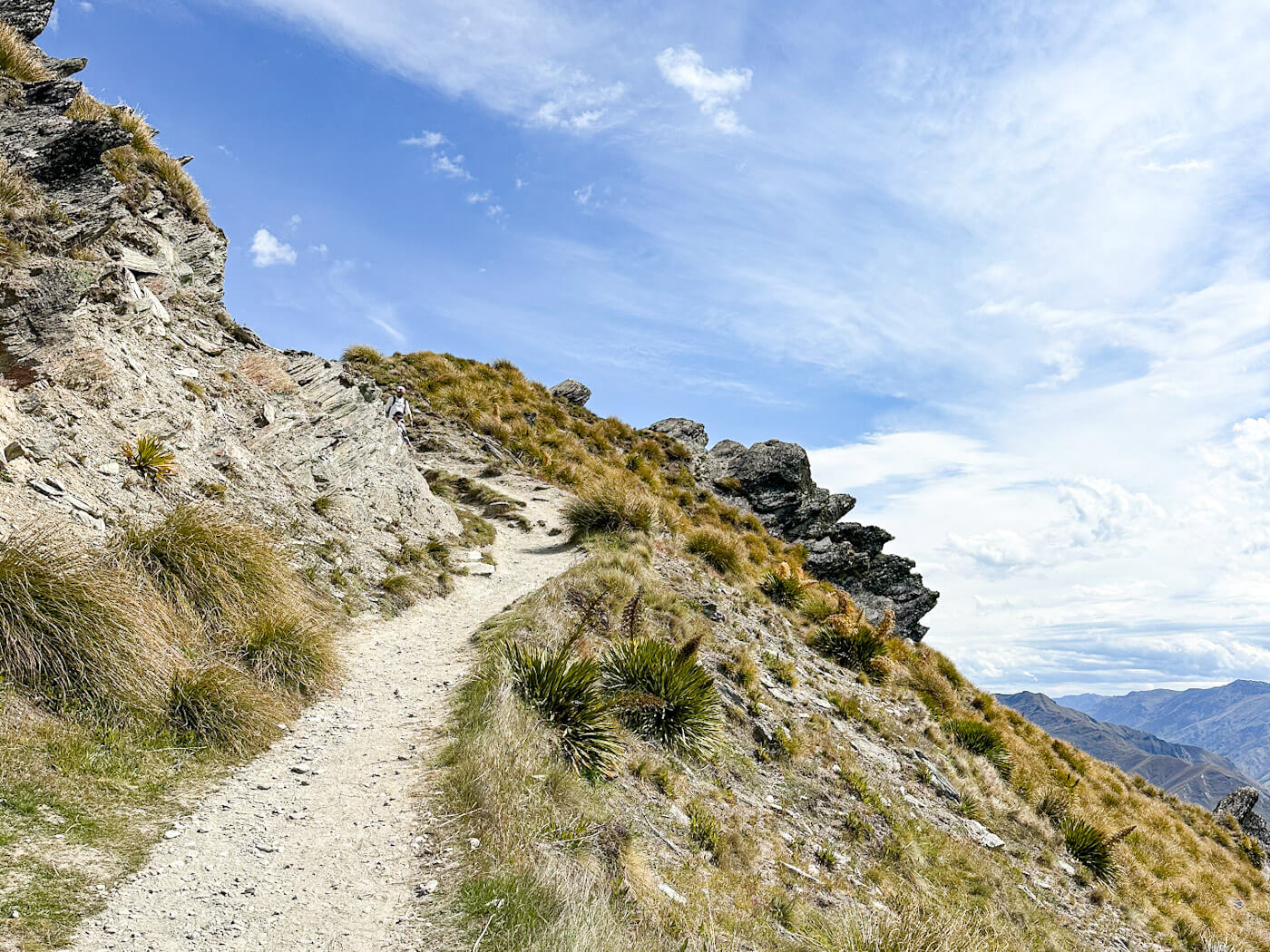 A perspective shot from a narrow, dirt hiking trail as it climbs steeply alongside a rugged, rocky cliff face. Sparse, golden-brown tussock grass grows in clumps along the dry, dusty incline. The trail leads upward toward a bright blue sky with wispy clouds, with a glimpse of distant blue mountains visible in the lower right corner.