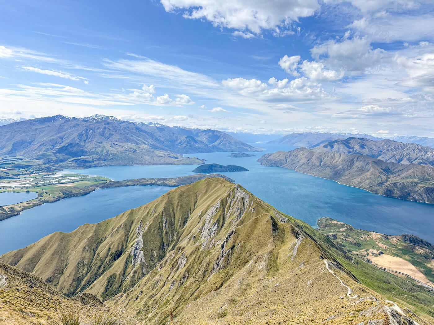 A breathtaking, wide-angle panorama from the very top of Roy's Peak. The foreground shows a steep, grassy mountain ridge descending into a sharp spine. Beyond, the vast, deep blue waters of Lake Wฤnaka are framed by golden-green hills and several small islands. On the horizon, a continuous chain of jagged, snow-capped mountains sits under a bright blue sky filled with scattered white clouds.