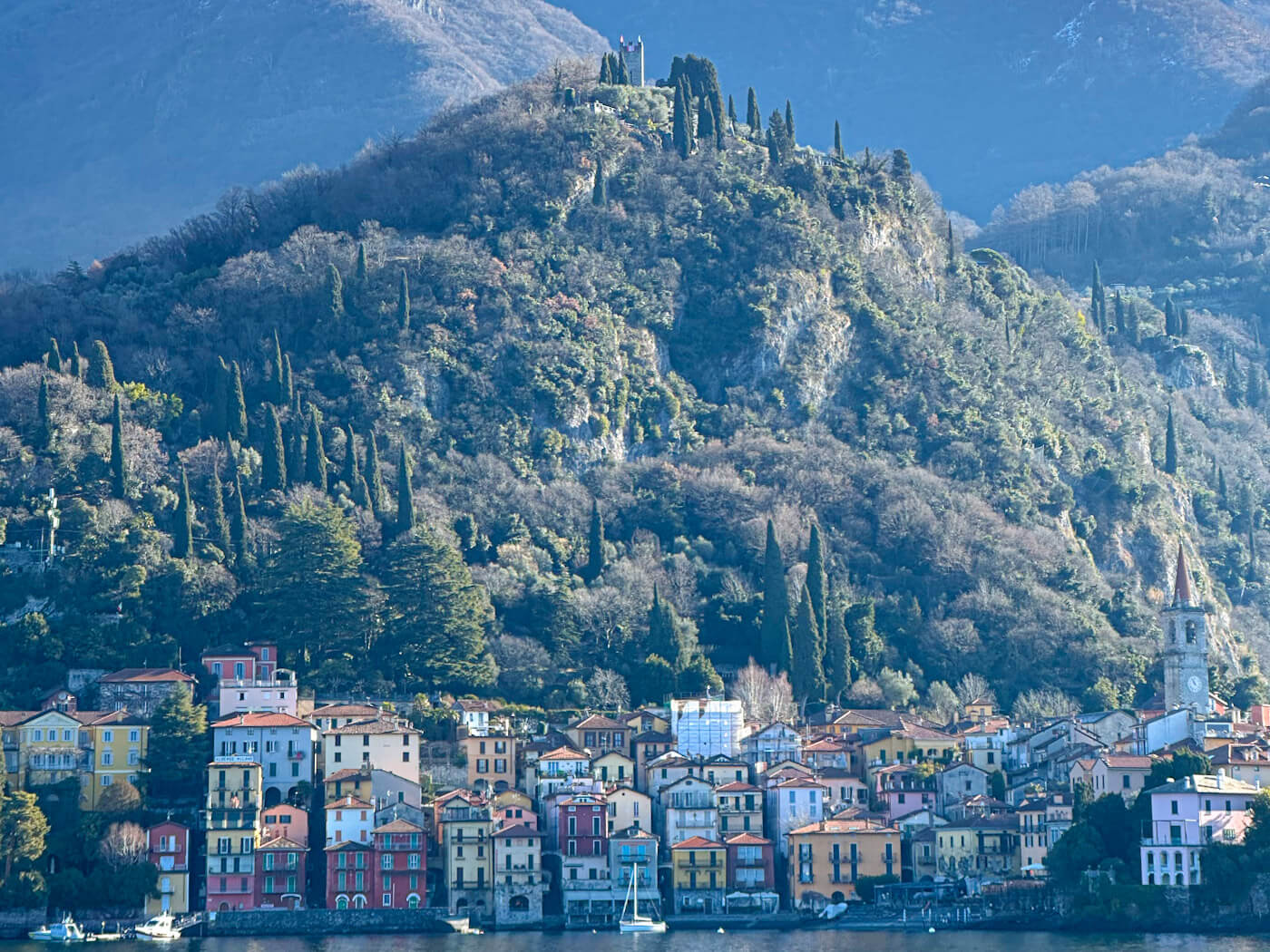 A picturesque, wide-angle view of the colorful waterfront village of Varenna on Lake Como, featuring a dense cluster of yellow, red, and peach buildings at the base of a steep, green mountain topped with a historic stone tower.