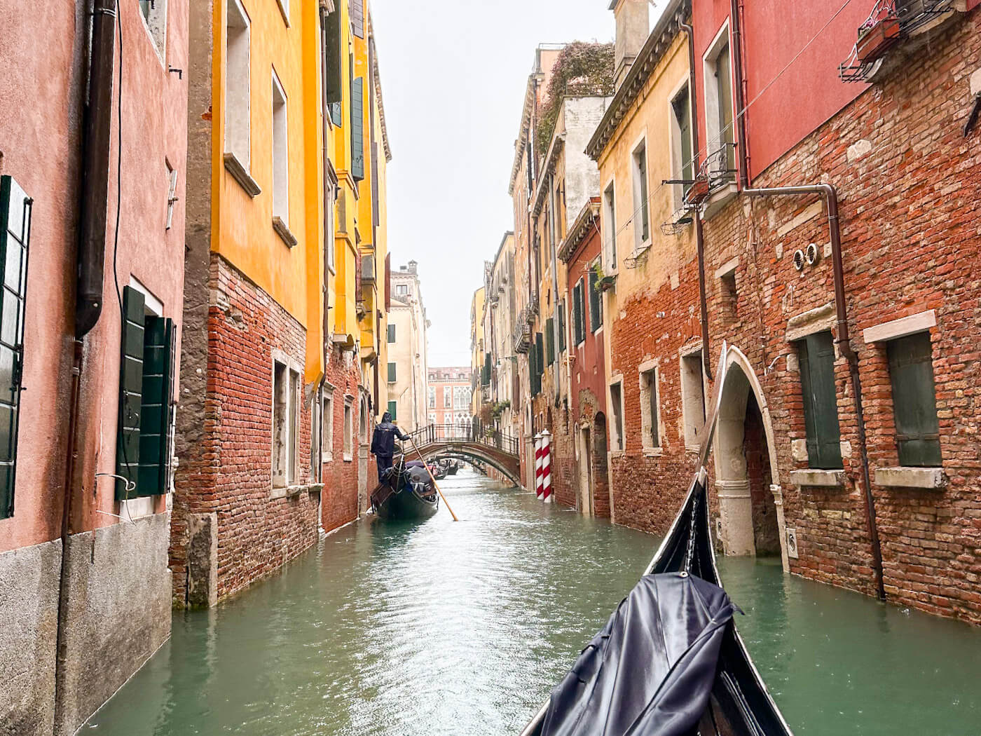 A first-person view from a gondola navigating a narrow waterway lined with tall, weathered brick buildings and passing under a distant footbridge in Venice in the winter.