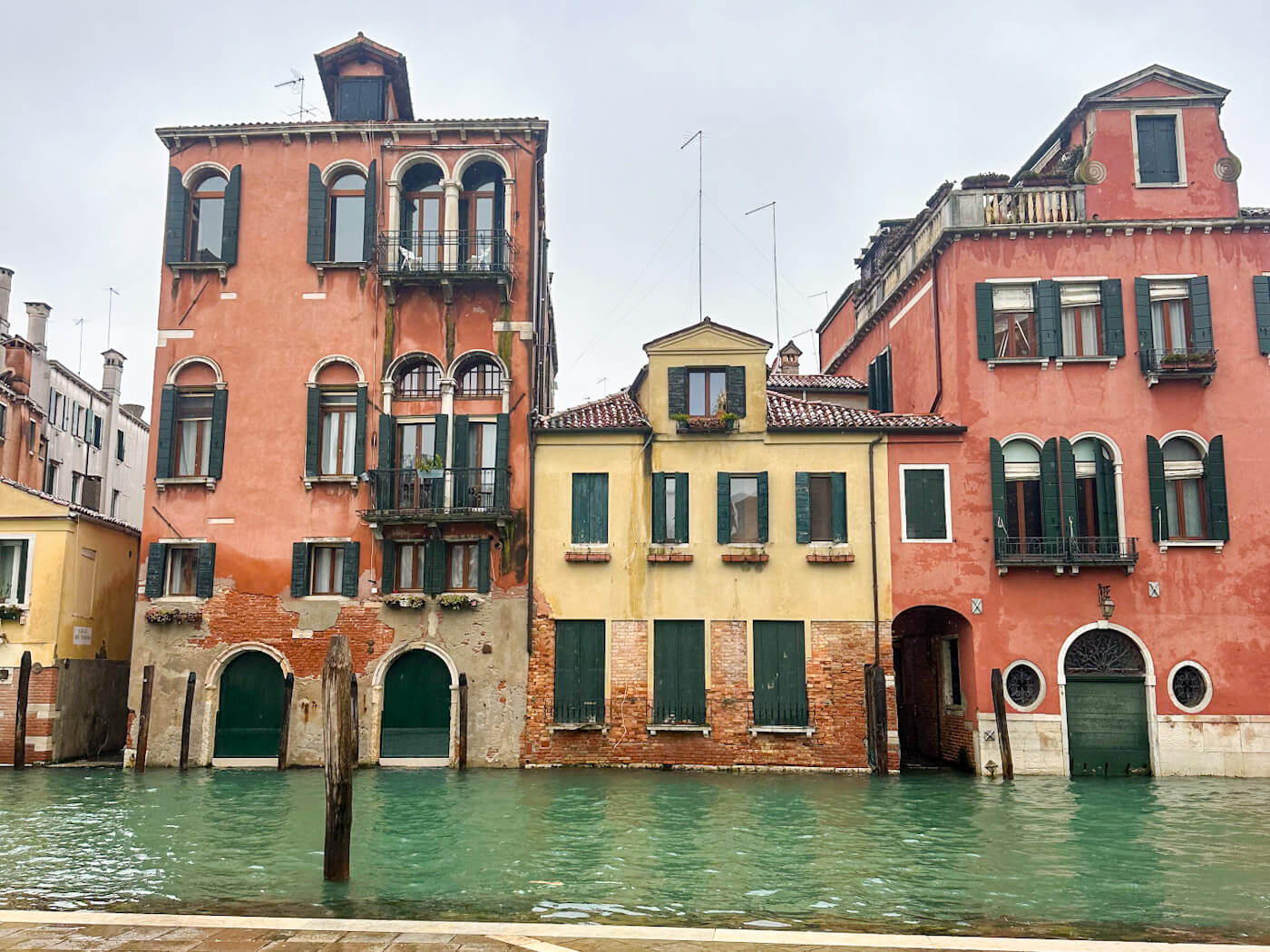A row of classic Venetian buildings in shades of peach and yellow with green shutters, situated directly on the turquoise water of the Grand Canal during a cloudy day in Venice in the winter.