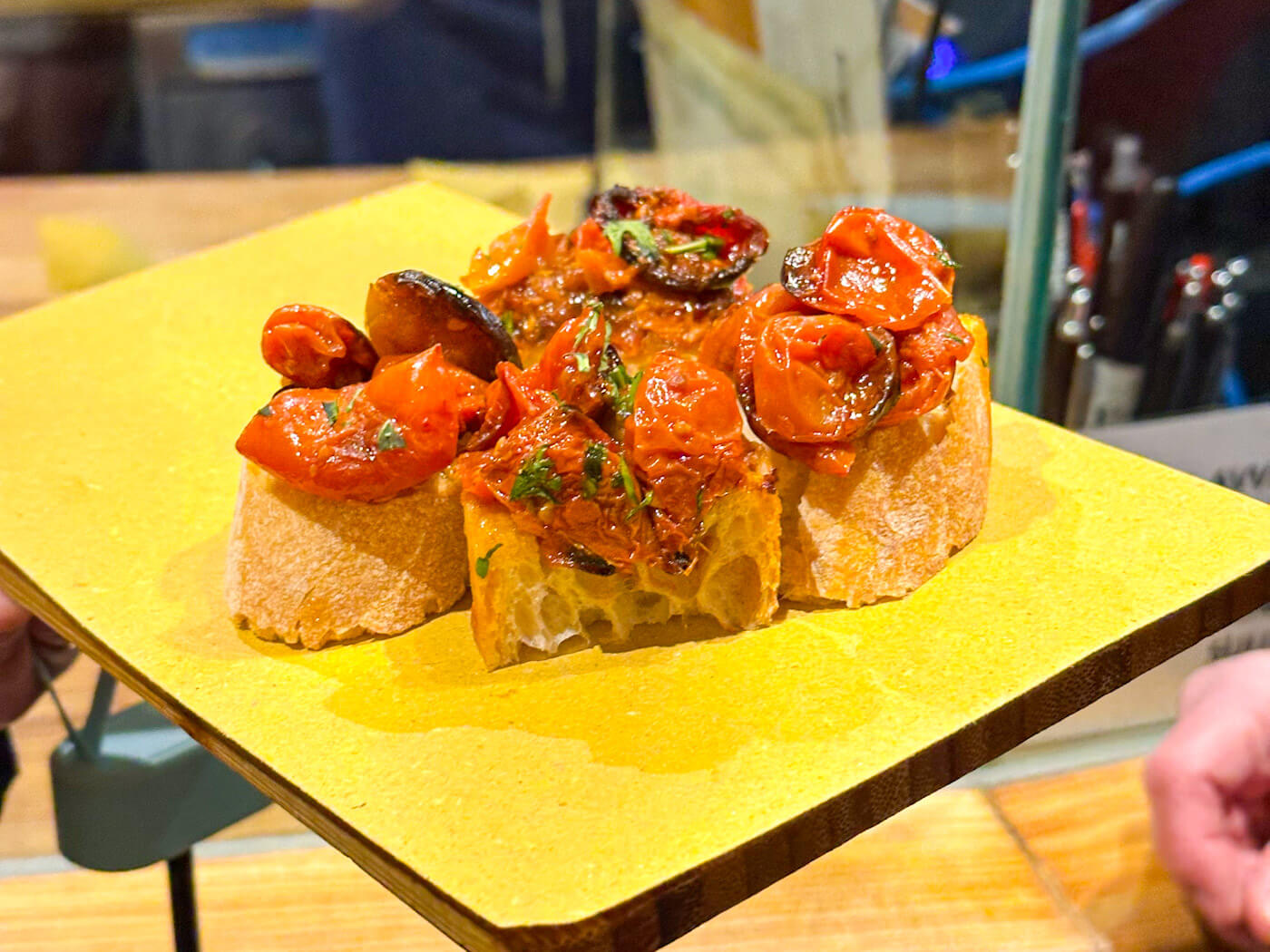 A close-up of Venetian cicchetti consisting of crusty bread topped with roasted balsamic tomatoes and herbs, served on a yellow napkin—a perfect warm snack while exploring Venice in the winter.