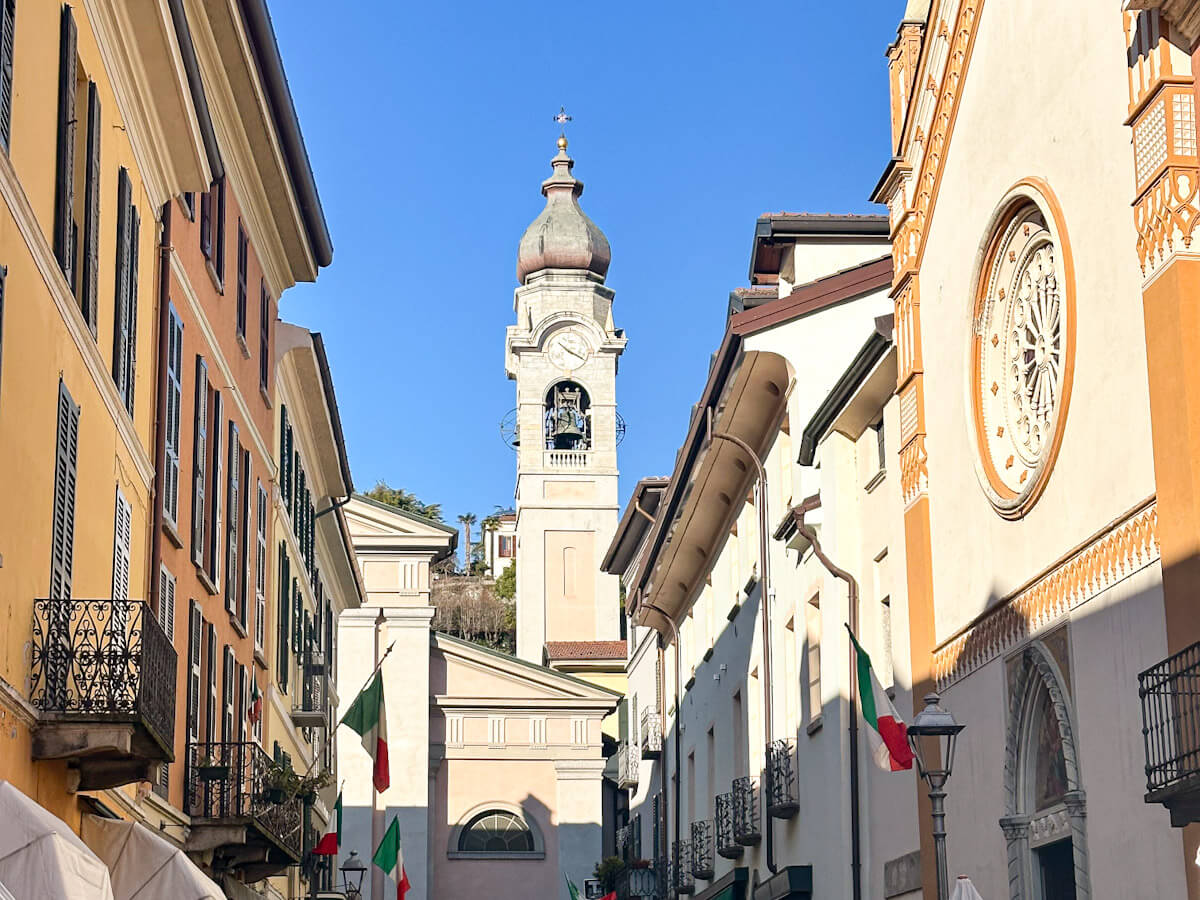 The bell tower of Chiesa di Santo Stefano rising above the cobbled streets of Menaggio, with Italian flags and colourful buildings lining the way — Menaggio is one of three charming towns to visit on a day trip to Lake Como