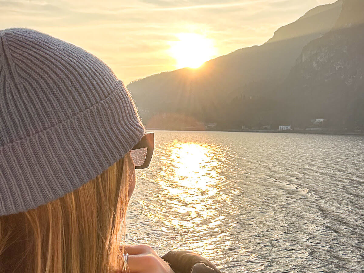 Charlie is wearing a blue knitted beanie and sunglasses and gazing out across the shimmering golden waters of Lake Como from the ferry deck, as the sun sets behind the dramatic mountain ridgeline — the kind of unforgettable moment that makes a day trip to Lake Como from Milan so worth it