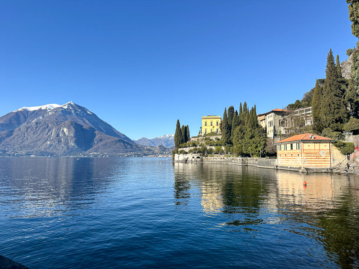 Crystal clear blue waters of Lake Como reflecting a grand yellow villa and tall cypress trees on the shoreline at Varenna, with snow-capped mountains stretching into the distance under a perfect blue sky