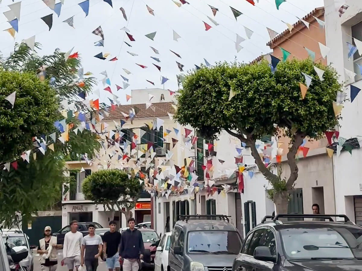 Colourful handmade triangular bunting flags strung across a sunny street in Es Castell during the Sant Jaume Menorca fiesta, with white-walled buildings and trimmed trees below