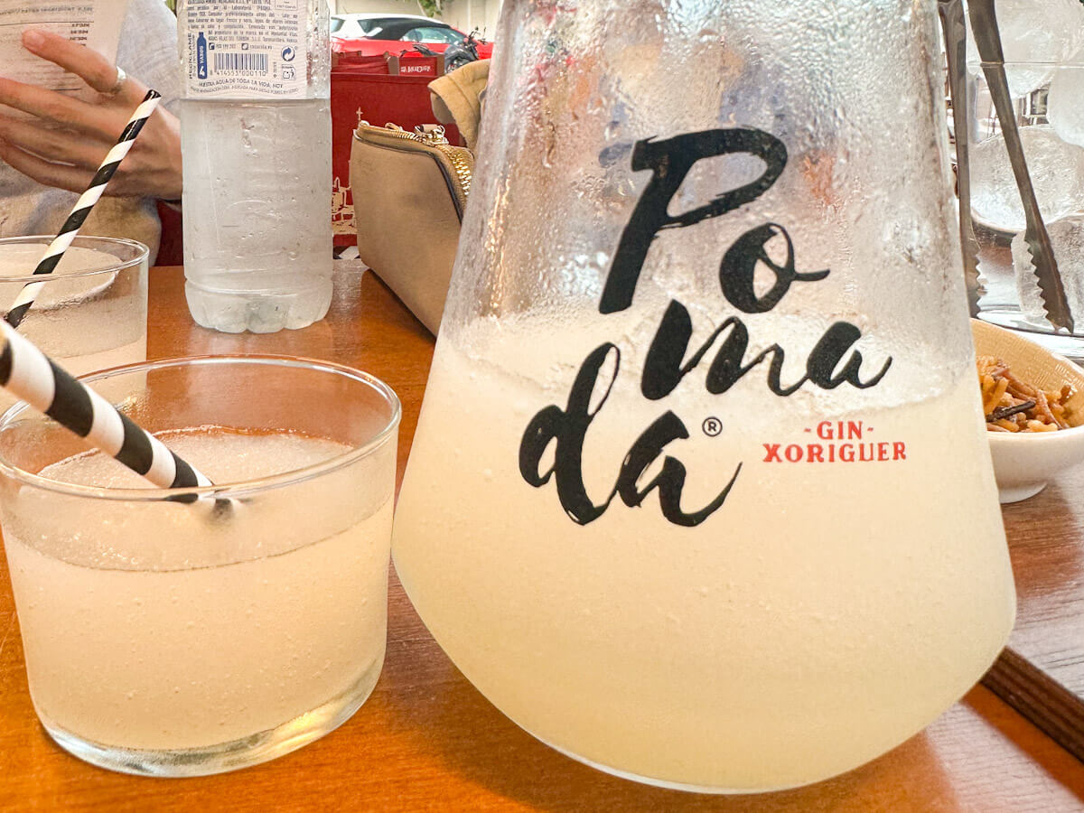 A chilled glass and pitcher of Pomada, a traditional Menorcan gin and lemonade cocktail, served with striped straws at a sun-drenched outdoor table