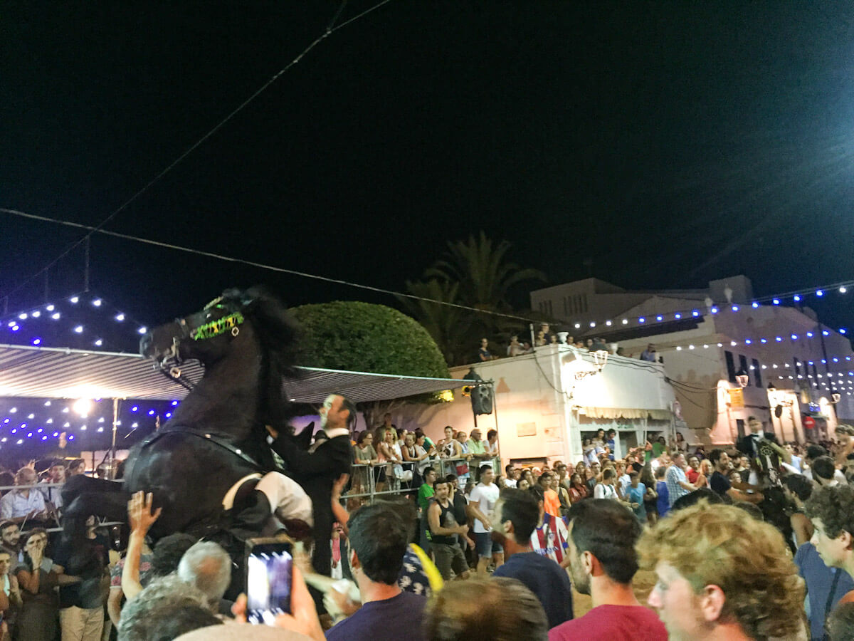 A black Menorquรญn horse rearing up dramatically during the Jaleo at the Sant Lluรญs Menorca fiesta, surrounded by a packed crowd of spectators in the main square at night