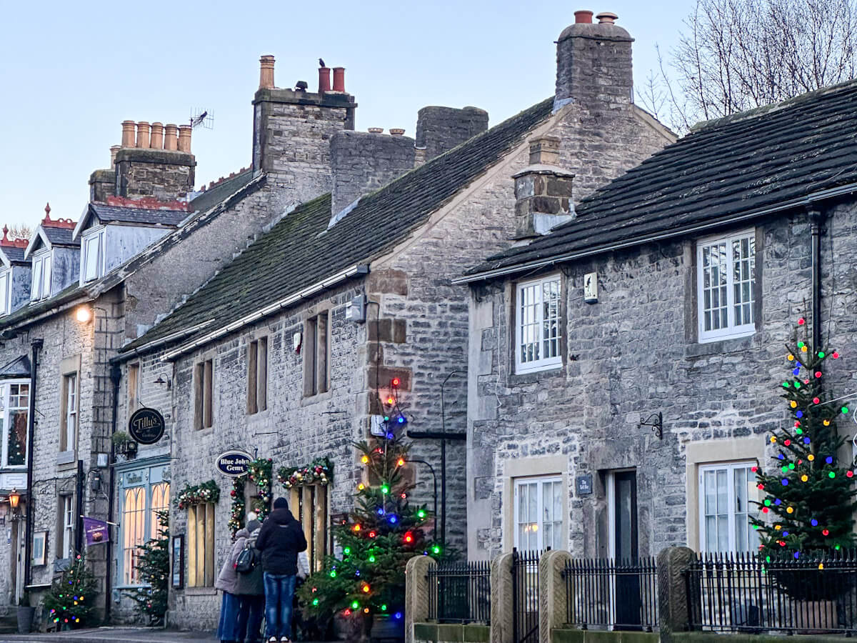 A row of traditional Derbyshire stone cottages in Castleton decorated for Christmas. Brightly colored holiday lights adorn several pine trees along the sidewalk as tourists walk past local shops like Tilly's of Castleton.