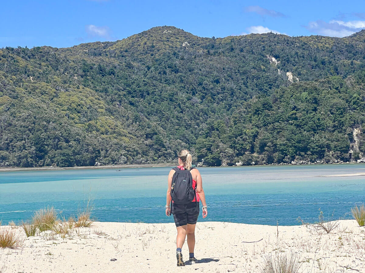 Charlie is wearing a black backpack walking along a wide golden sand beach towards large turquoise tidal flats and a large, forest-covered hill in Abel Tasman National Park.