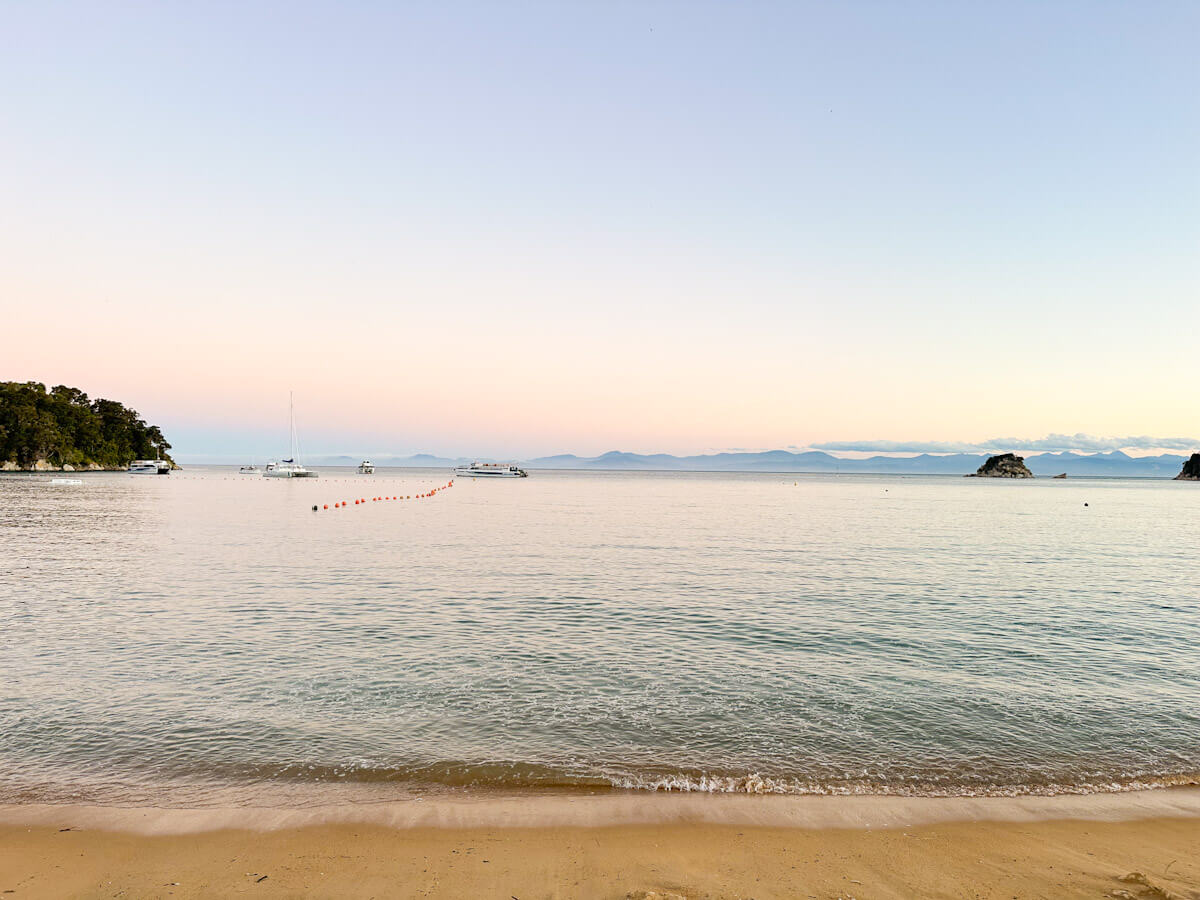 A calm, soft pastel sunset over the iconic golden sands of Kaiteriteri Beach, with several boats and a water taxi anchored in the tranquil bay, a key hub for things to do in Abel Tasman.