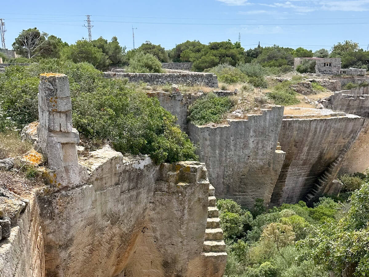 A high-angle view of the Lithica quarry showing ancient stone structures, rock-cut stairs, and lush green vegetation growing within the excavated pits.