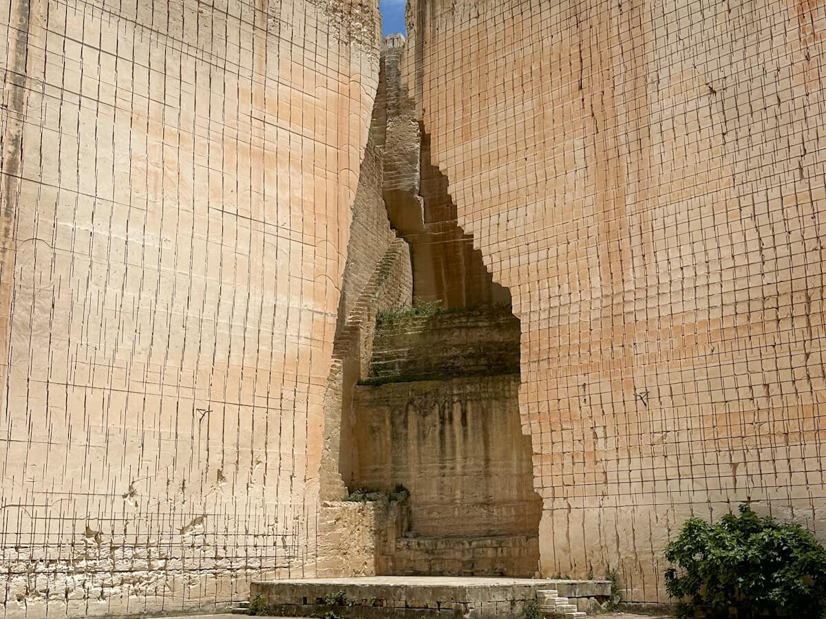 A view looking up at the towering, sheer vertical sandstone walls of the Lithica quarry, featuring precise grid-like cutting marks from historic stone extraction.