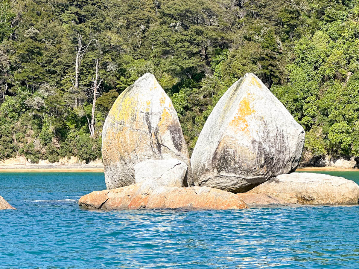 A detailed close-up shot of the famous Split Apple Rock, a large granite boulder split perfectly in two, resting on orange-tinted rocks in blue water near the coastline.
