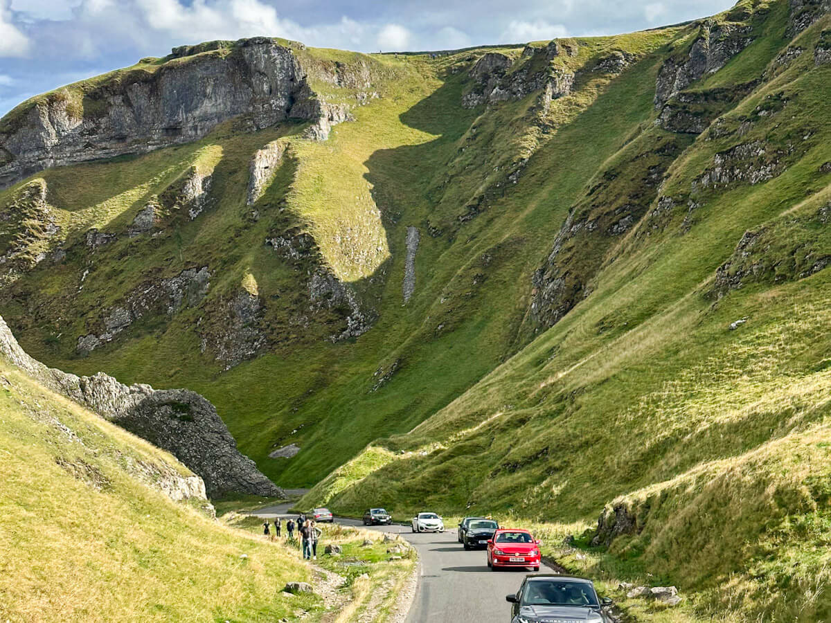 A winding road snakes through the dramatic, steep limestone cliffs of Winnats Pass near Castleton. Several cars drive through the lush green gorge while a group of hikers stands on the grass by the roadside.