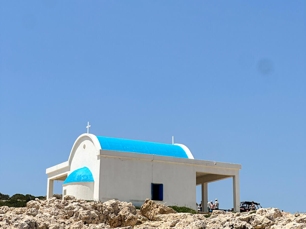 A classic white Greek Orthodox chapel with a vibrant, bright blue domed roof standing on a rocky coastline against a clear blue sky in Cape Greco, Cyprus.