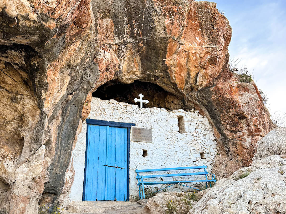 The unique Agioi Saranta cave church built into a rocky hillside near Ayia Napa, Cyprus, featuring a bright blue wooden door, a matching blue bench, and a simple white cross above the entrance.