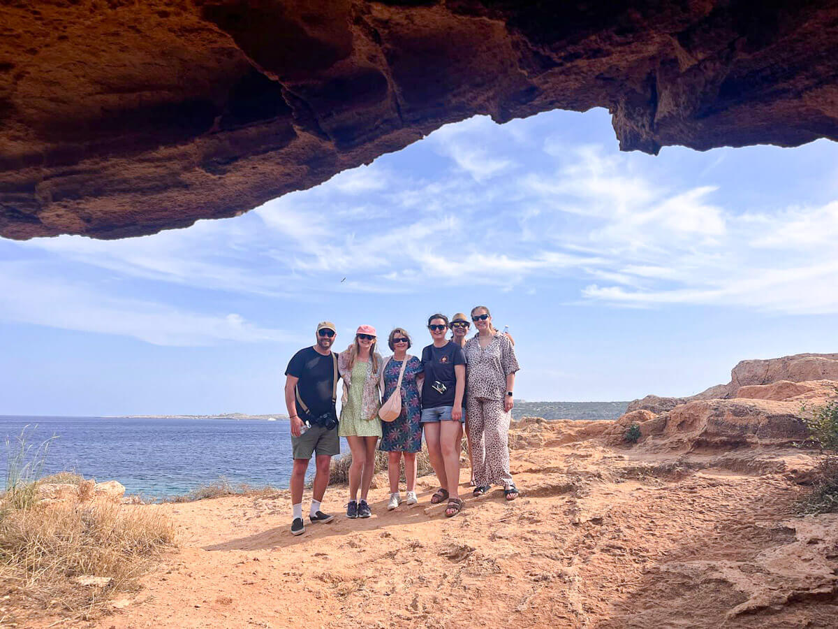A group of travelers posing together on a dusty trail overlooking the blue Mediterranean Sea, framed from inside the dark opening of Cyclops Cave in Cape Greco near Ayia Napa.