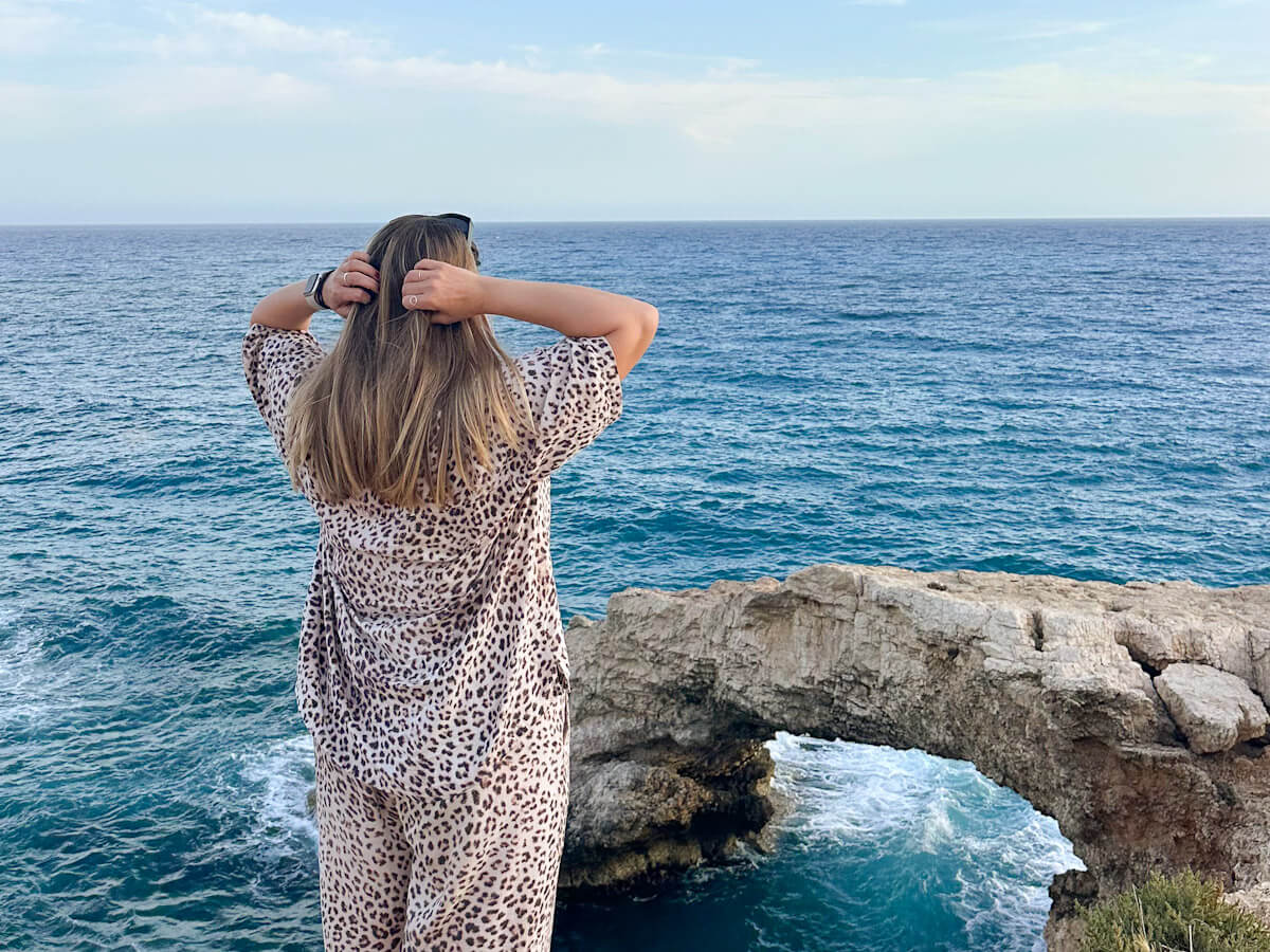 Charlie from behind, wearing a leopard-print outfit and fixing her hair, while looking out over the natural stone archway of Love Bridge and the deep blue Mediterranean Sea in Ayia Napa. What to do in Ayia Napa