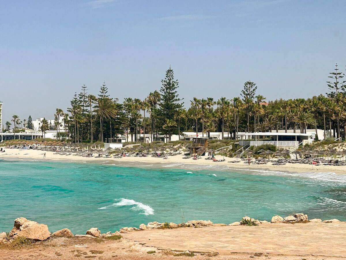 The turquoise waters and white sands of Nissi Beach, lined with sun loungers, palm trees, and beachfront resorts under a hazy blue sky.