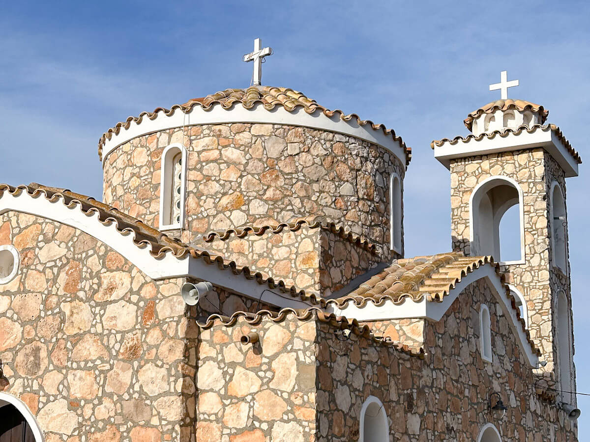 A close-up view of the historic Profitis Ilias stone church in Protaras near Ayia Napa, showcasing its traditional stone-brick architecture, arched windows, terracotta roof tiles, and two white stone crosses against a soft blue sky. What to do in Ayia Napa