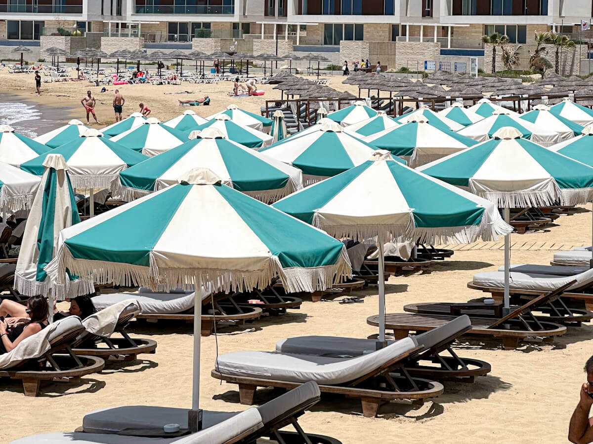 Rows of distinctive green and white striped umbrellas with fringed edges shading wooden sun loungers on the sandy beach at Riva Beachouse in Ayia Napa Marina, with modern beachfront accommodation in the background. What to do in Ayia Napa