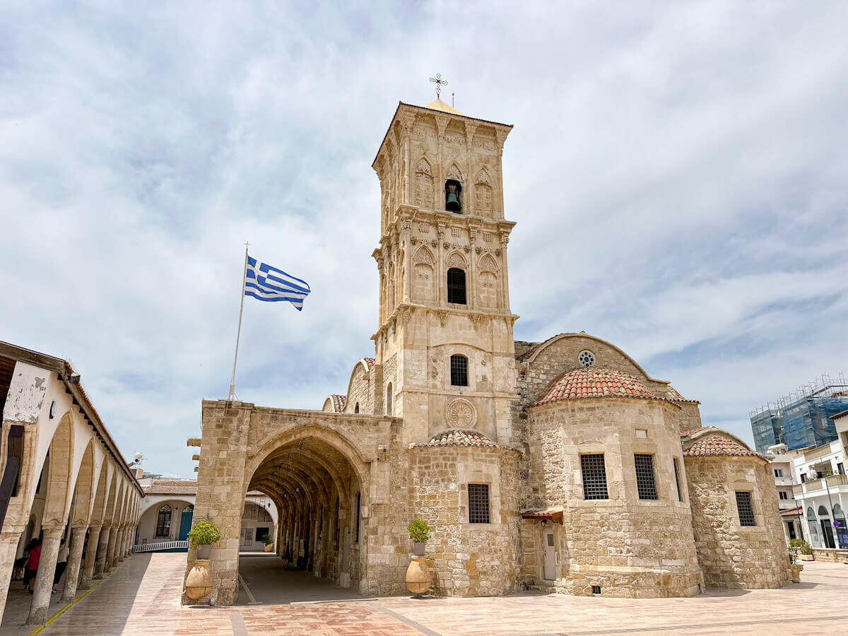 The historic stone architecture and tall bell tower of the Church of Saint Lazarus under a cloudy sky, with a Greek flag flying in the plaza—a top choice for what to do in Larnaca.