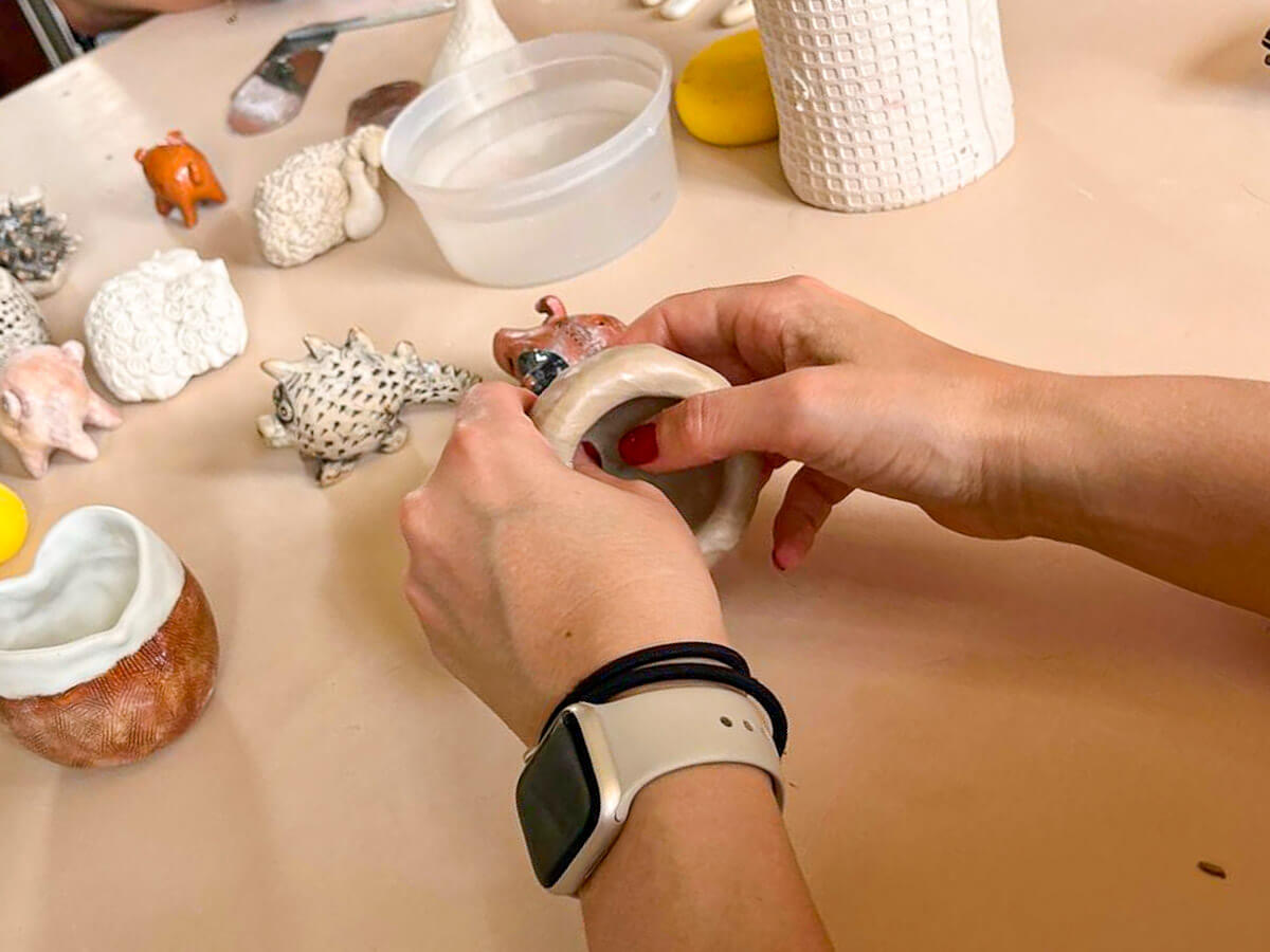 A close-up of a person's hands sculpting a small clay bowl at a table during a studio ceramics pottery class, surrounded by handmade clay animals and crafting tools, highlighting a unique option for what to do in Larnaca.