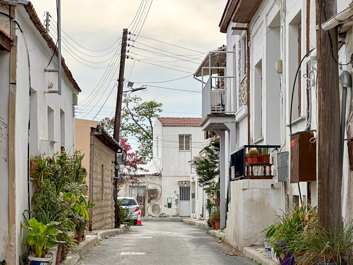 A quiet, narrow street in the historic Turkish Cypriot quarter, featuring traditional white-walled buildings, potted green plants, and overhead power lines, highlighting what to do in Larnaca.