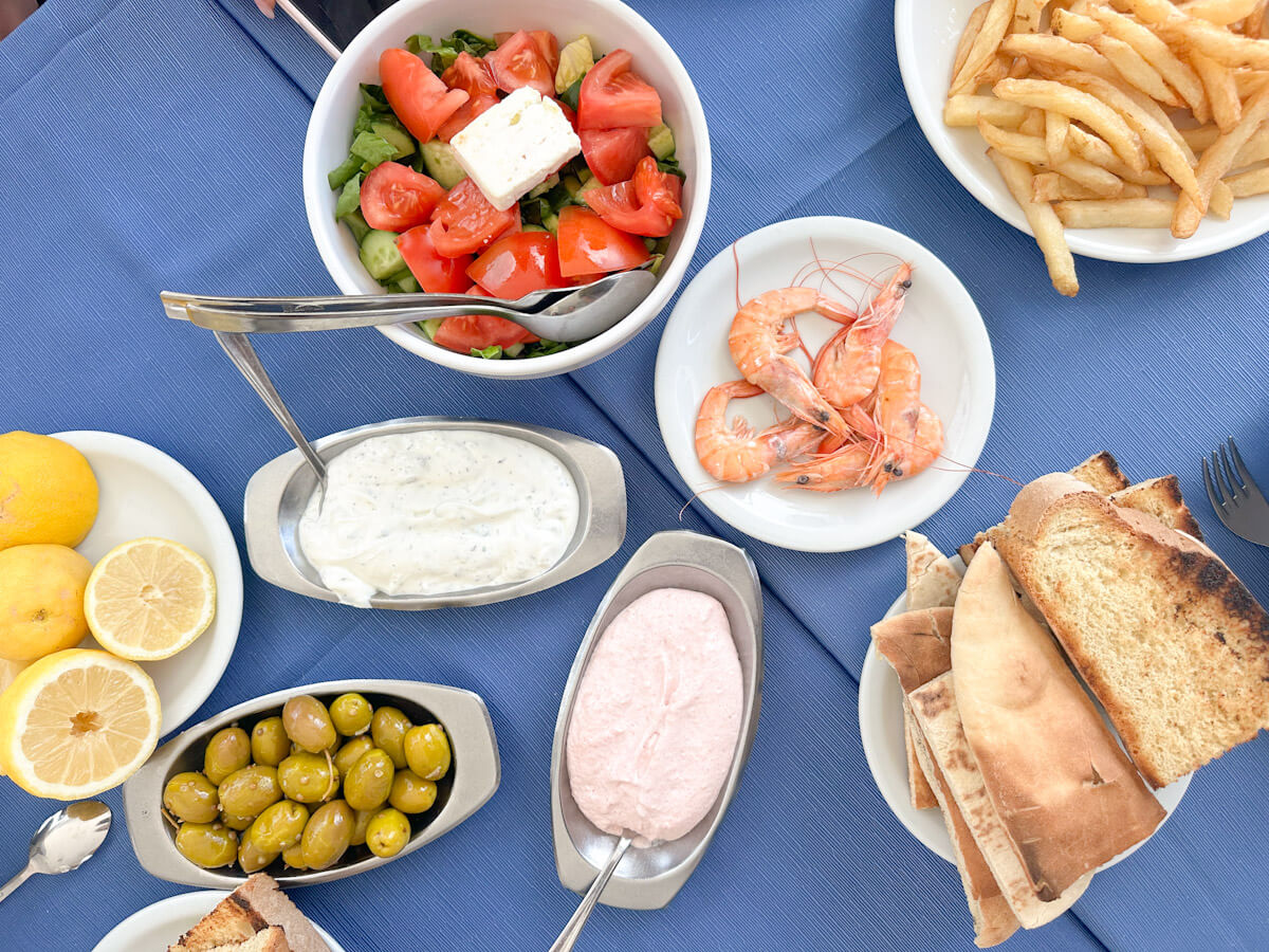 A top-down view of a traditional Cypriot meze spread on a blue tablecloth at Zephyros Restaurant, featuring a Greek salad with feta cheese, green olives, pita and toasted bread, French fries, prawns, lemons, and bowls of tzatziki and taramasalata dips.