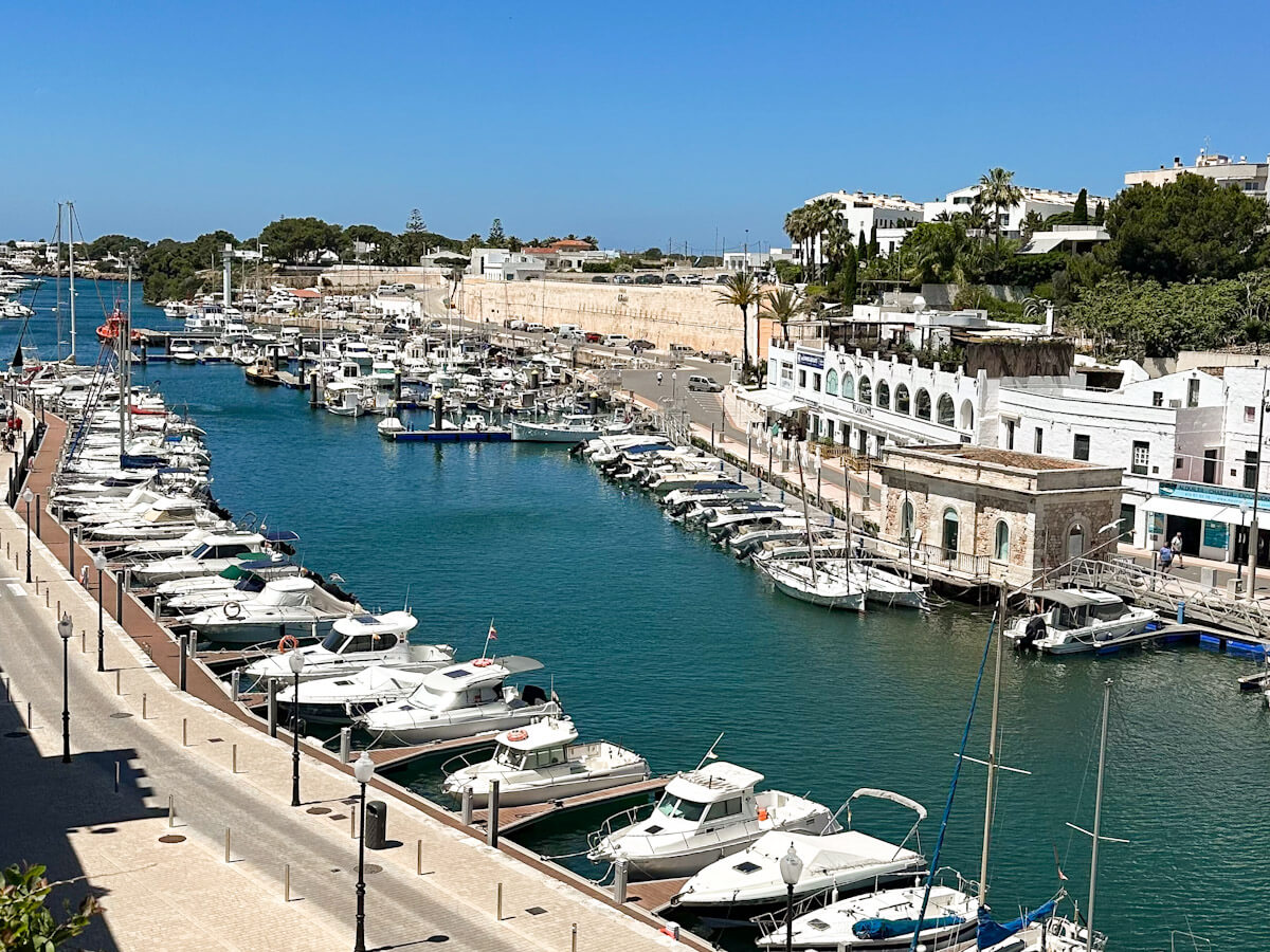 A panoramic view of the bustling marina in Ciutadella, Menorca, filled with numerous boats docked in turquoise water, lined by stone buildings and a promenade under a clear blue sky