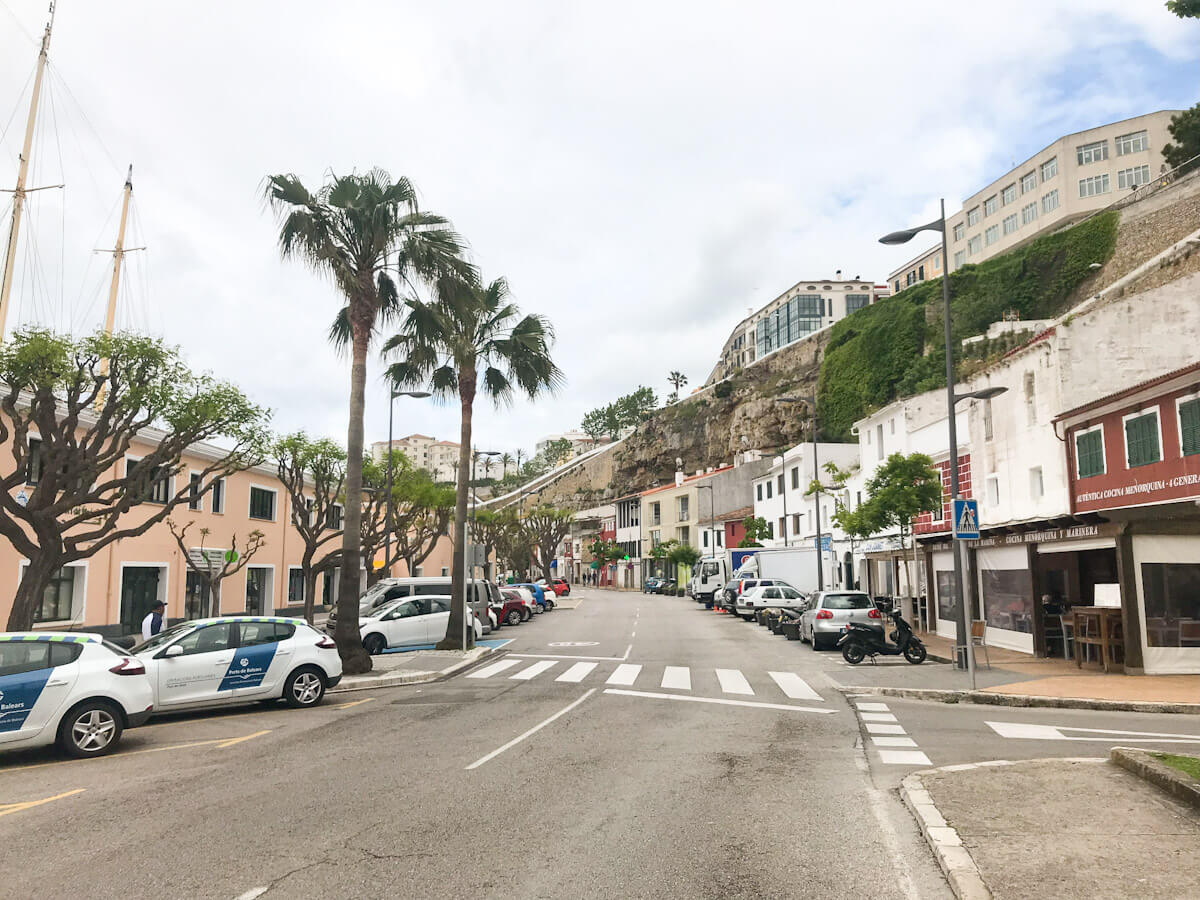 A wide shot of the picturesque Moll de Llevant waterfront area, showing palm trees, parked cars, and traditional coastal buildings nestled against a tall, rocky cliffsideโa scenic stop for anyone exploring things to do in Mahon.
