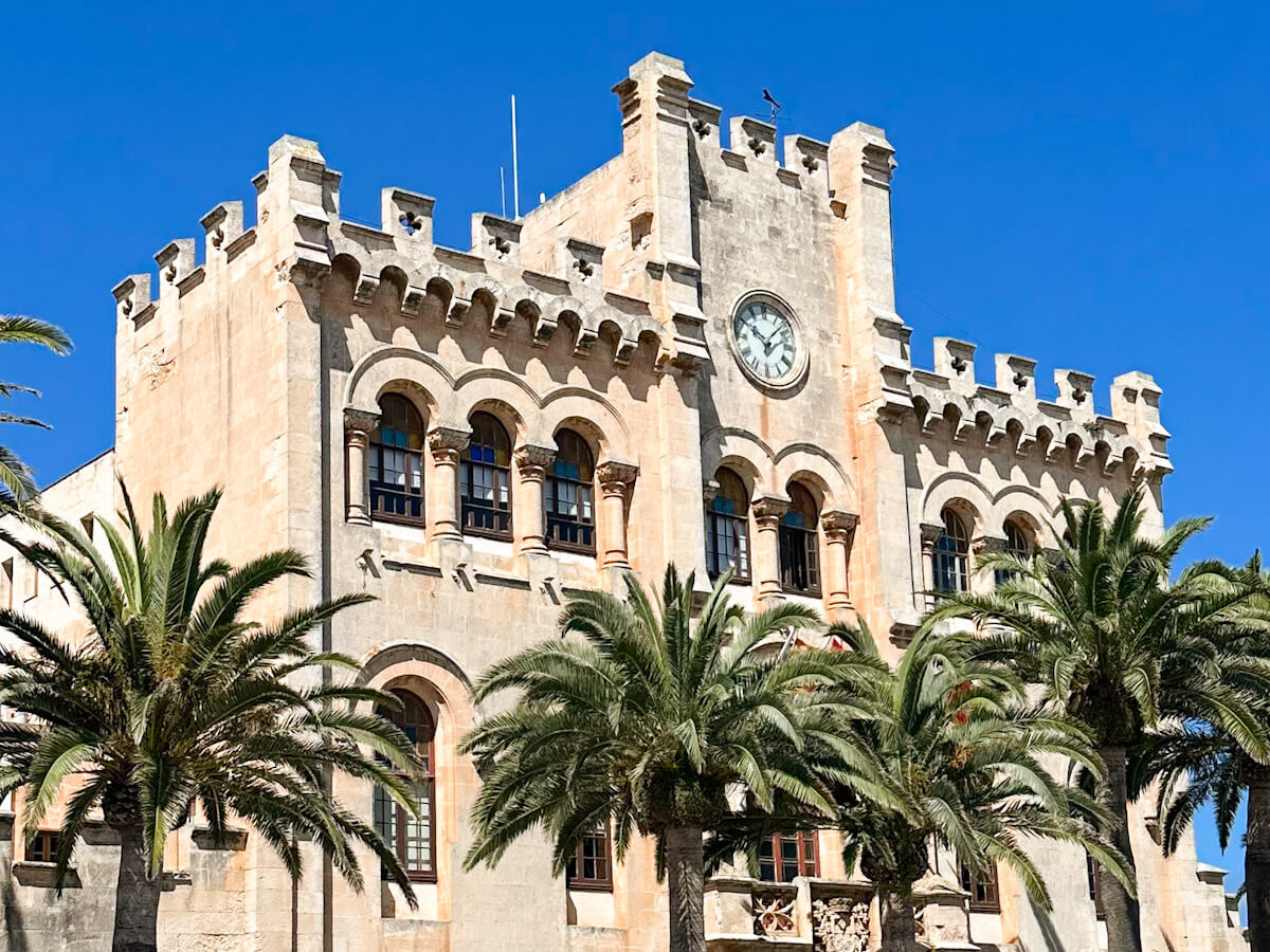 An architectural detail of the historic City Hall building in Plaça des Born, Ciutadella, showcasing its crenelated roofline, a prominent clock, and lush palm trees in the foreground.