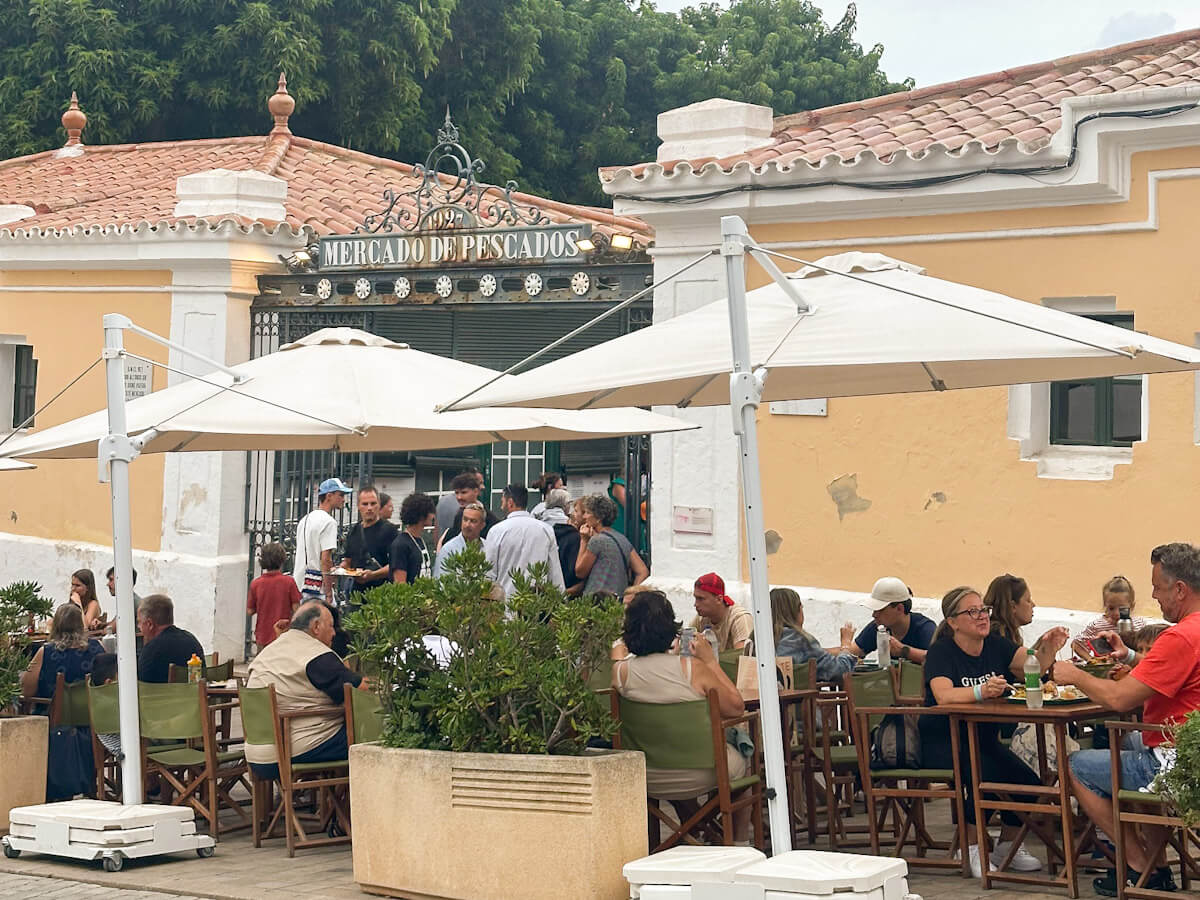 A lively scene with diners sitting at outdoor cafe tables under umbrellas in front of the historic stone building of the Mercat des Peix, highlighting a classic food-focused experience among things to do in Mahon.