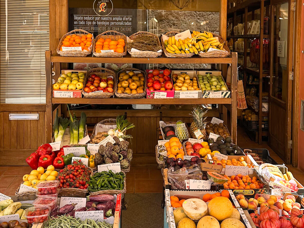 A vibrant display of fresh fruits and vegetables, including oranges, bananas, tomatoes, and peppers, arranged in wooden crates and baskets inside a municipal market.
