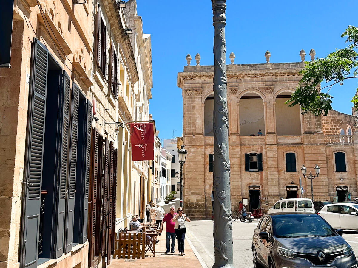 A sunny street in Ciutadella, Menorca, featuring traditional historic buildings with dark wooden shutters, a decorative stone lamppost in the foreground, and people walking on the sidewalk