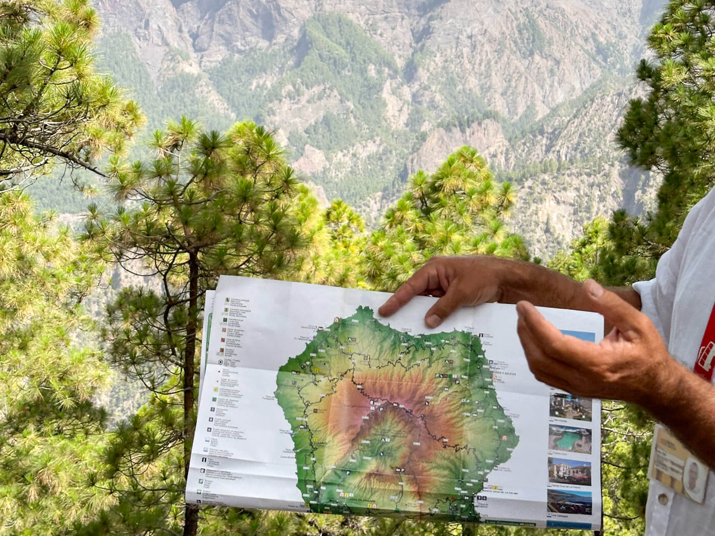 A person holding up a topographic map of La Palma with lush pine trees and the dramatic, sweeping canyon depths of Caldera de Taburiente National Park in the background, highlighting hiking planning as a key part of things to do in La Palma.