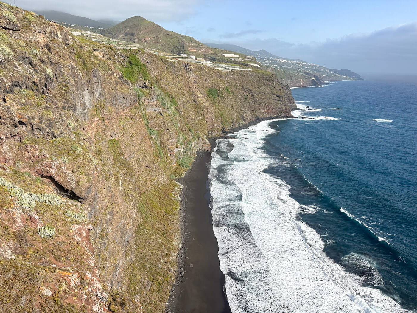 High-angle view of the dramatic black sand beach and crashing white waves at Playa de Nogales in Puntallana against towering green cliffs, a visual highlight among things to do in La Palma.