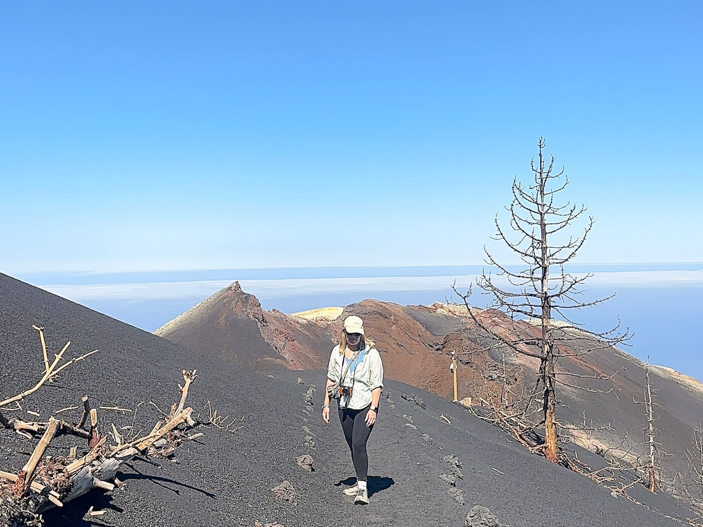 Charlie is walking along a black volcanic ash path with a skeletal burnt tree and the tajogaite volcano in the background, capturing outdoor exploration as one of the top things to do in La Palma.