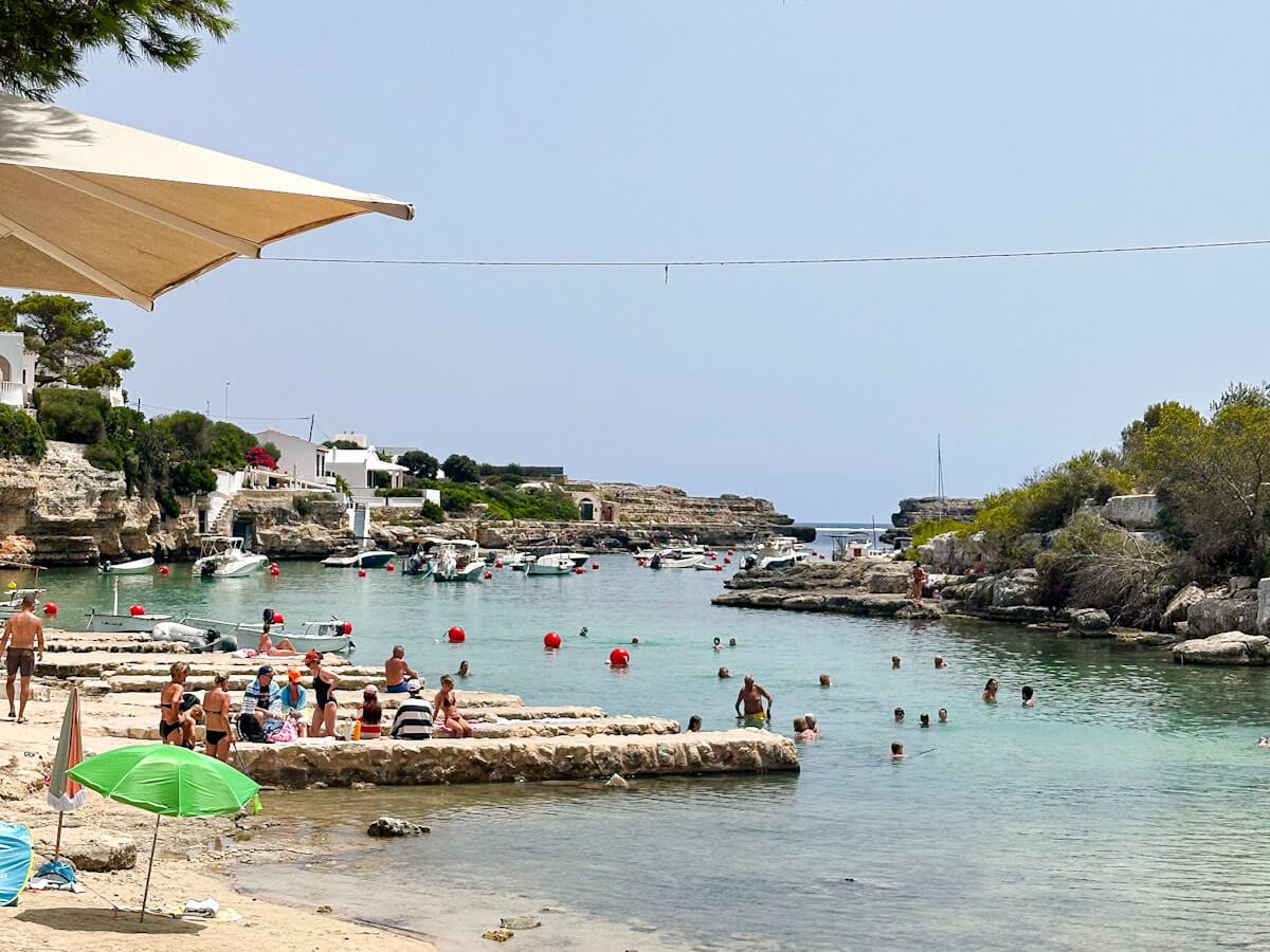 Swimmers relaxing on stone platforms and wading in the shallow, clear turquoise waters of the rocky cove of Alcaufar in Menorca, with small boats anchored in the background under a clear sky. Ideal when visiting Menorca in September
