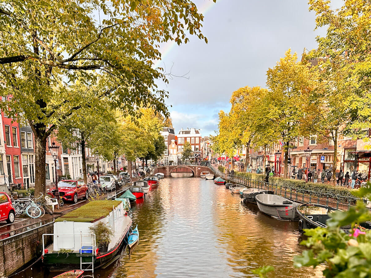 A scenic view of an Amsterdam canal lined with autumn trees, moored boats, and historic buildings under a soft sky with a faint rainbow.