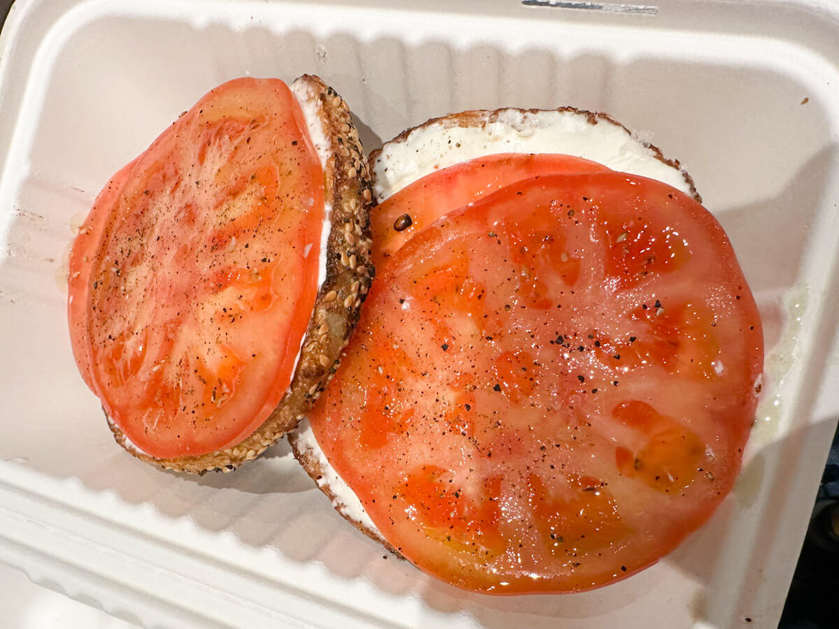 An open-faced Apollo Bagel topped with a layer of cream cheese and thick slices of ripe tomato seasoned with black pepper, served in a takeout container as part of the best bagels in New York.