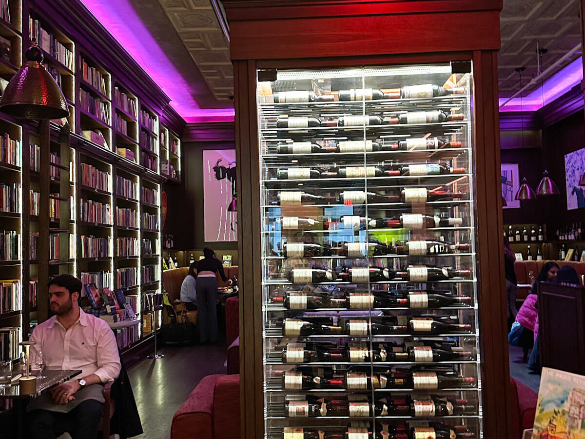 The interior of Bibliothéque in New York featuring floor-to-ceiling bookshelves, purple lighting accents, and a large illuminated glass wine display case in the centre, highlighting the cosy bar atmosphere for food in new york.