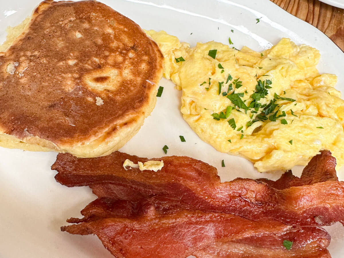 A classic weekend plate at Bubby's featuring a golden brown pancake, fluffy scrambled eggs garnished with herbs, and crispy bacon strips, making it a staple for the best brunch in new york as part of any food in new york journey.
