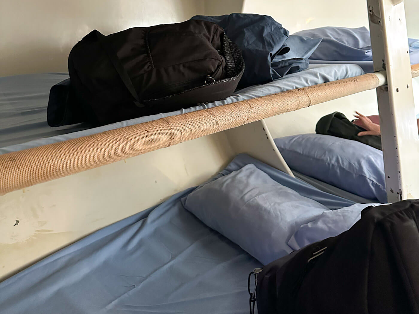 A close-up view of narrow bunk beds inside a sailboat cabin, outfitted with light blue sheets and pillows, with a black duffel bag resting on the upper bunk during a trip sailing the Whitsundays.