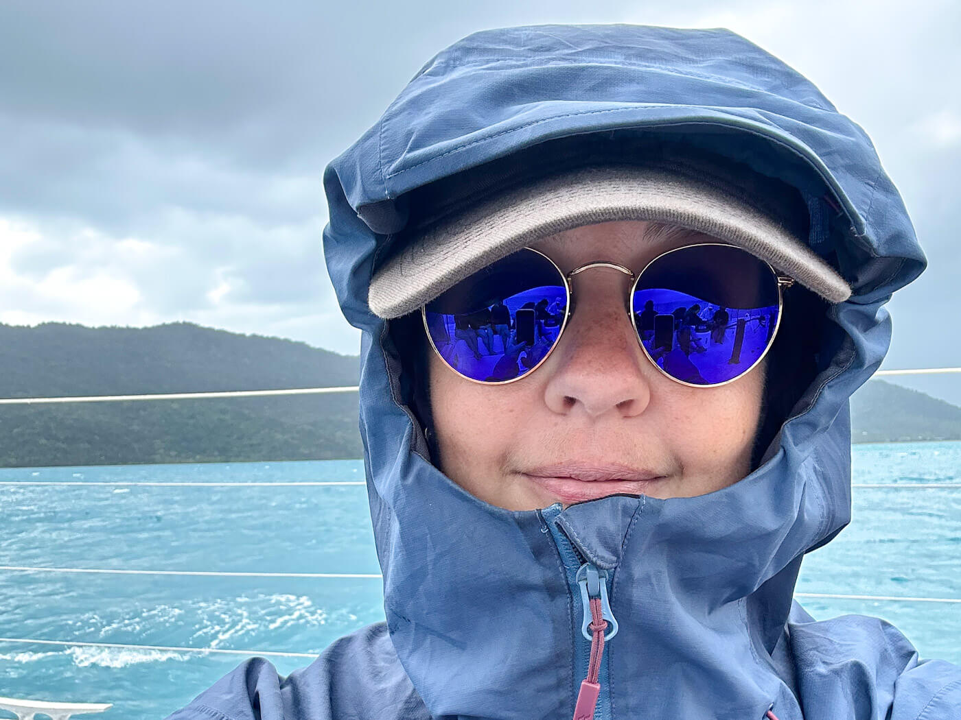 A close-up selfie of Charlie bundled up in a blue waterproof jacket with the hood up and reflective round sunglasses, standing on a sailboat deck with blue water and islands under a cloudy sky while sailing the Whitsundays.