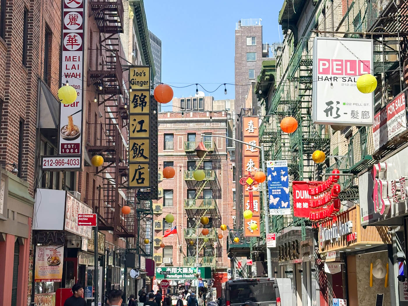 A bustling New York City Chinatown street view filled with historic brick buildings featuring fire escapes, vibrant restaurant signs in English and Chinese characters, and strings of glowing yellow and orange lanterns overhead, capturing the cultural scene for food in New York.