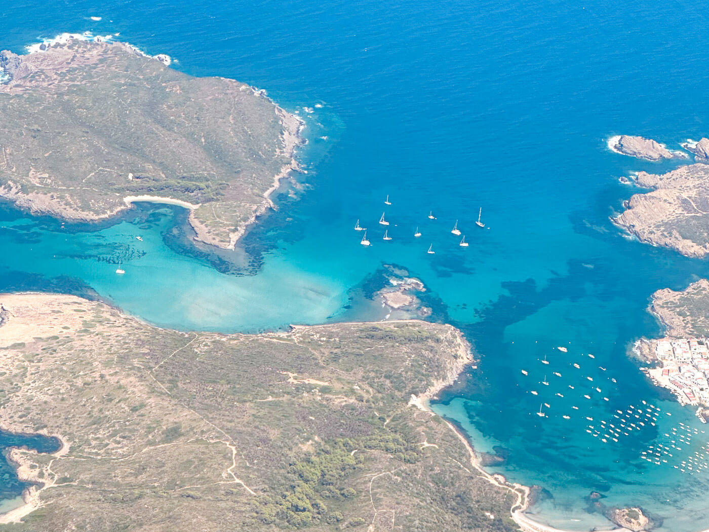 Aerial view of Menorca's coastline from a flight, showing vibrant turquoise waters, rocky green headlands, and sailboats anchored in the bay near a coastal town.