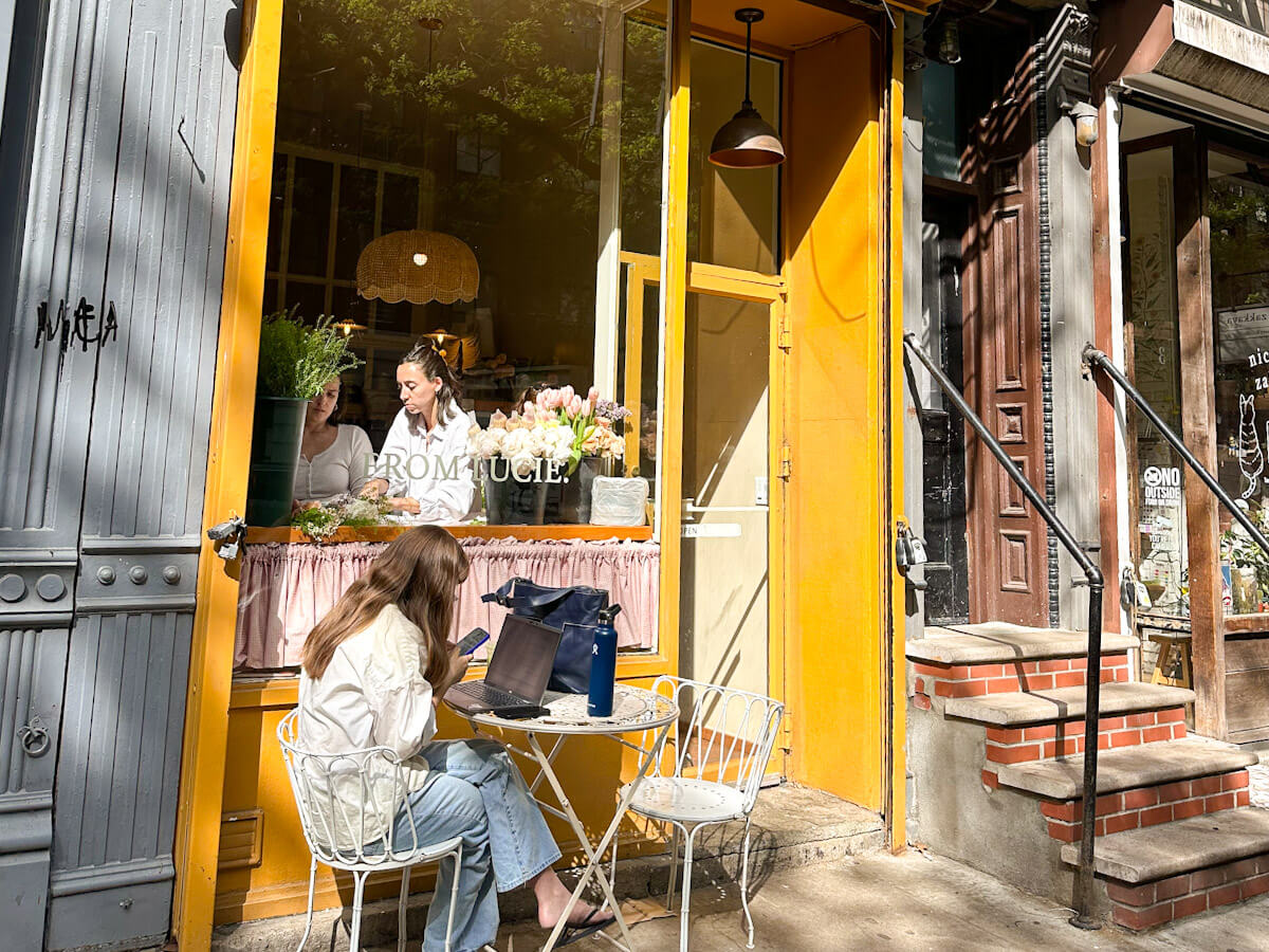 A sunny exterior view of the bright yellow storefront of From Lucie bakery in the East Village, with workers arranging fresh flowers inside the large window and a woman sitting outside at a white bistro table using her laptop and phone, representing the vibrant cafe scene for food in new york.